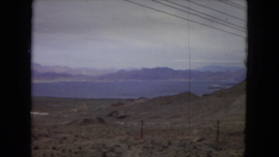 1967 United States 8mm frame showing scrubland, low desert ridges, a distant lake or reservoir, layered mountains, fence posts, and diagonal utility wires.