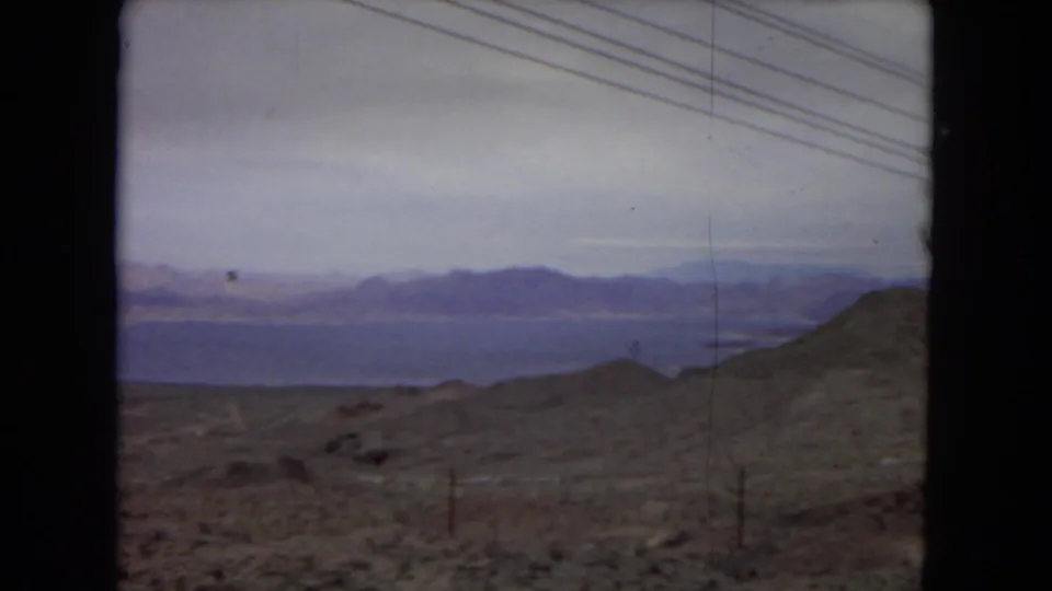 1967 United States home-movie frame of open desert terrain, fence posts, distant water, layered mountain ranges, and diagonal power lines under a hazy sky.