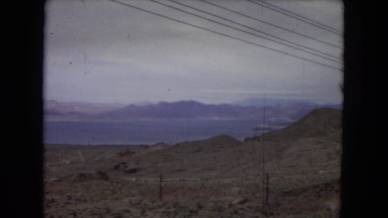 1967 8mm home-movie view from inside a moving vehicle in the United States, looking across arid hills toward distant water and mountains under a cloudy sky.