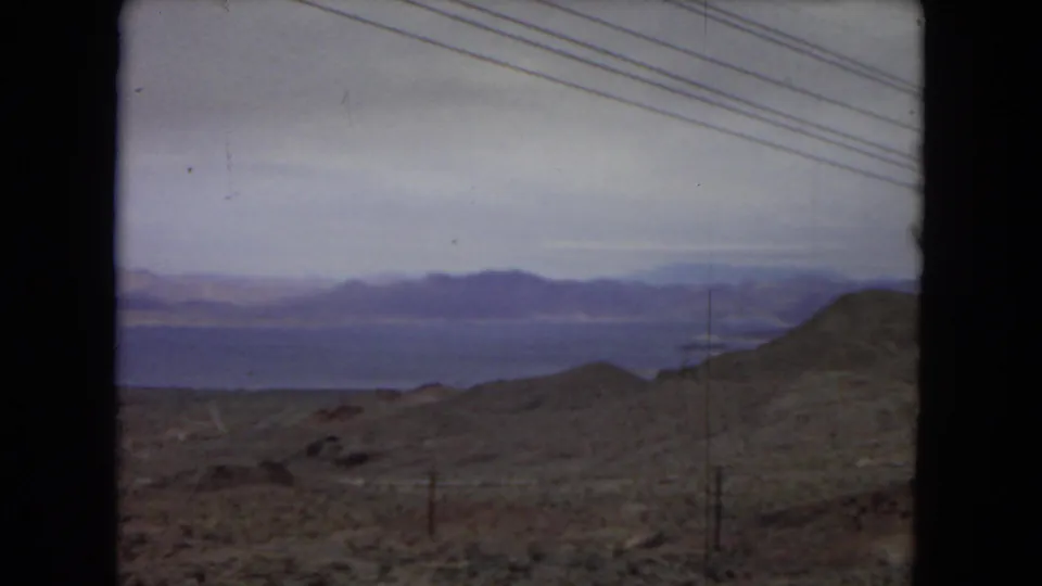 1967 8mm home-movie view from inside a moving vehicle in the United States, looking across arid hills toward distant water and mountains under a cloudy sky.