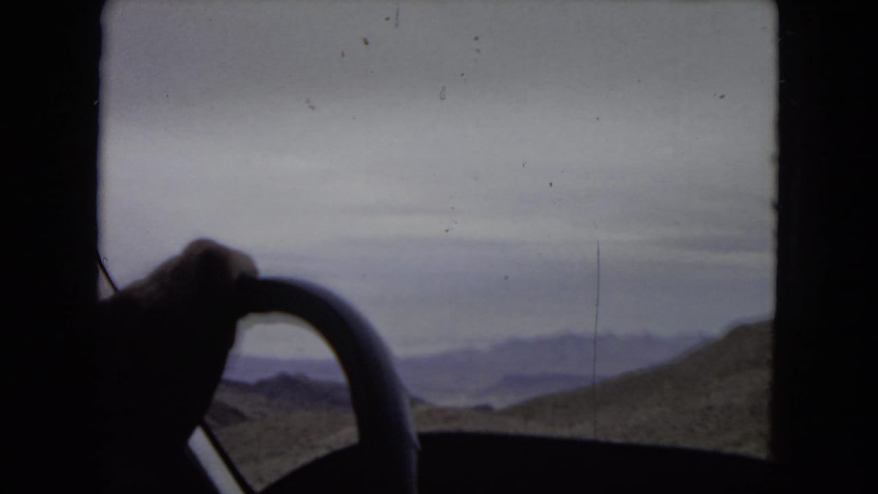 Interior driving view from 1967 home-movie footage, with a hand on the steering wheel silhouetted against desert mountains, water, and cloudy sky.