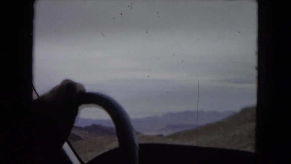 Interior driving view from 1967 home-movie footage, with a hand on the steering wheel silhouetted against desert mountains, water, and cloudy sky.