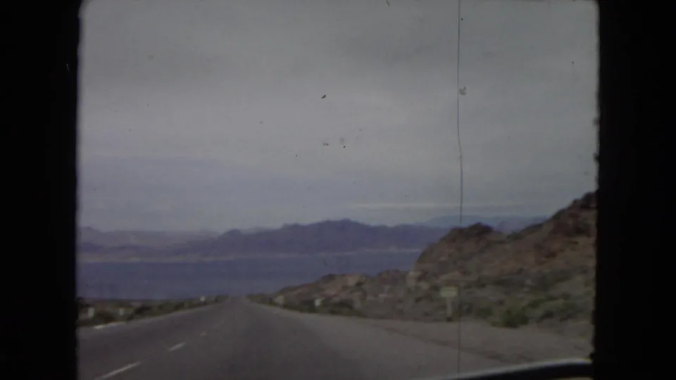 1967 8mm travel footage shows a paved desert highway running through rocky terrain toward distant mountains and a broad body of water.