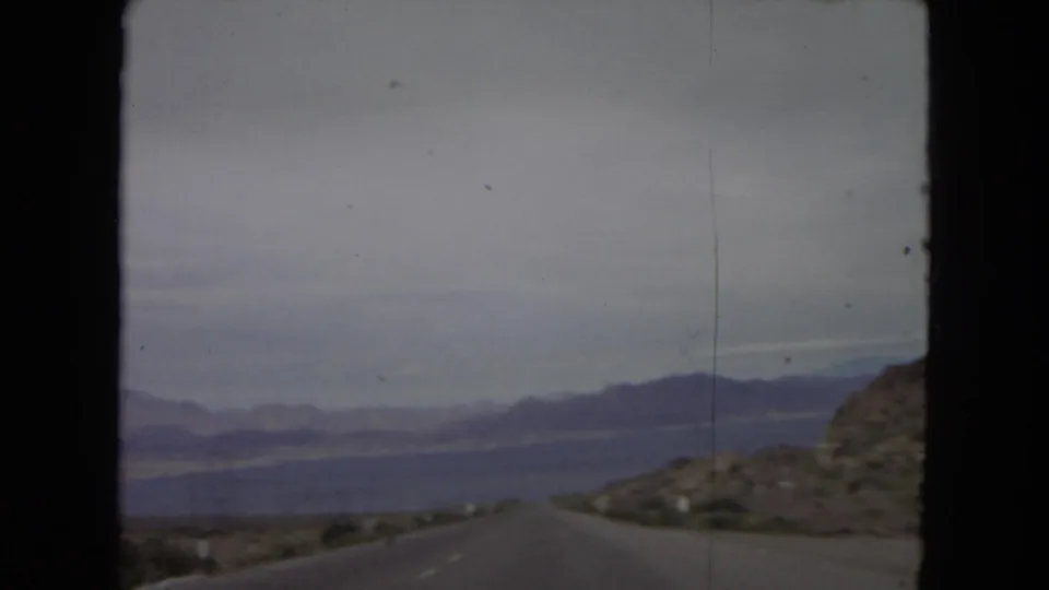 End frame of the 1967 8mm road-travel clip, with the highway leading through arid hills toward water, mountains, and a muted horizon.