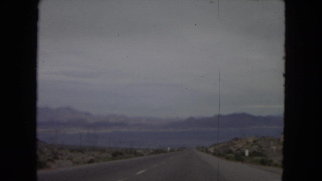 1967 8mm road-trip frame showing a long desert highway from inside a moving car, with distant mountains and a lake visible ahead.
