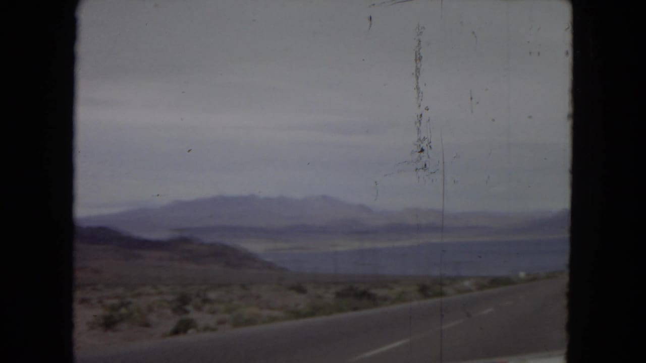Desert road curving beside a lake with mountains across the water.