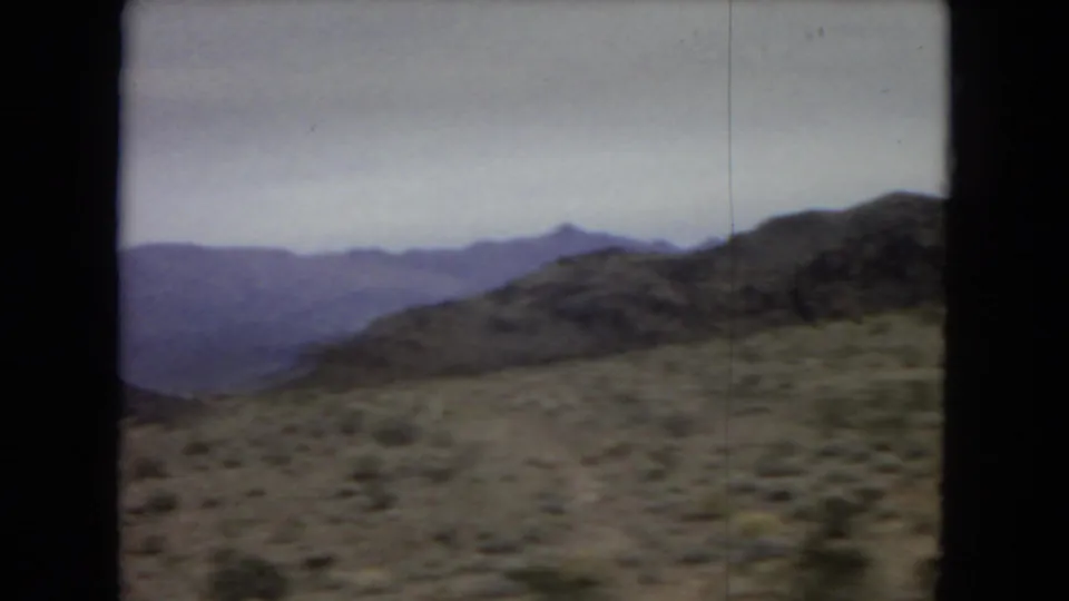 1967 8mm home-movie frame showing scrubby desert terrain and hazy mountain ridges viewed through a car window in the western United States.