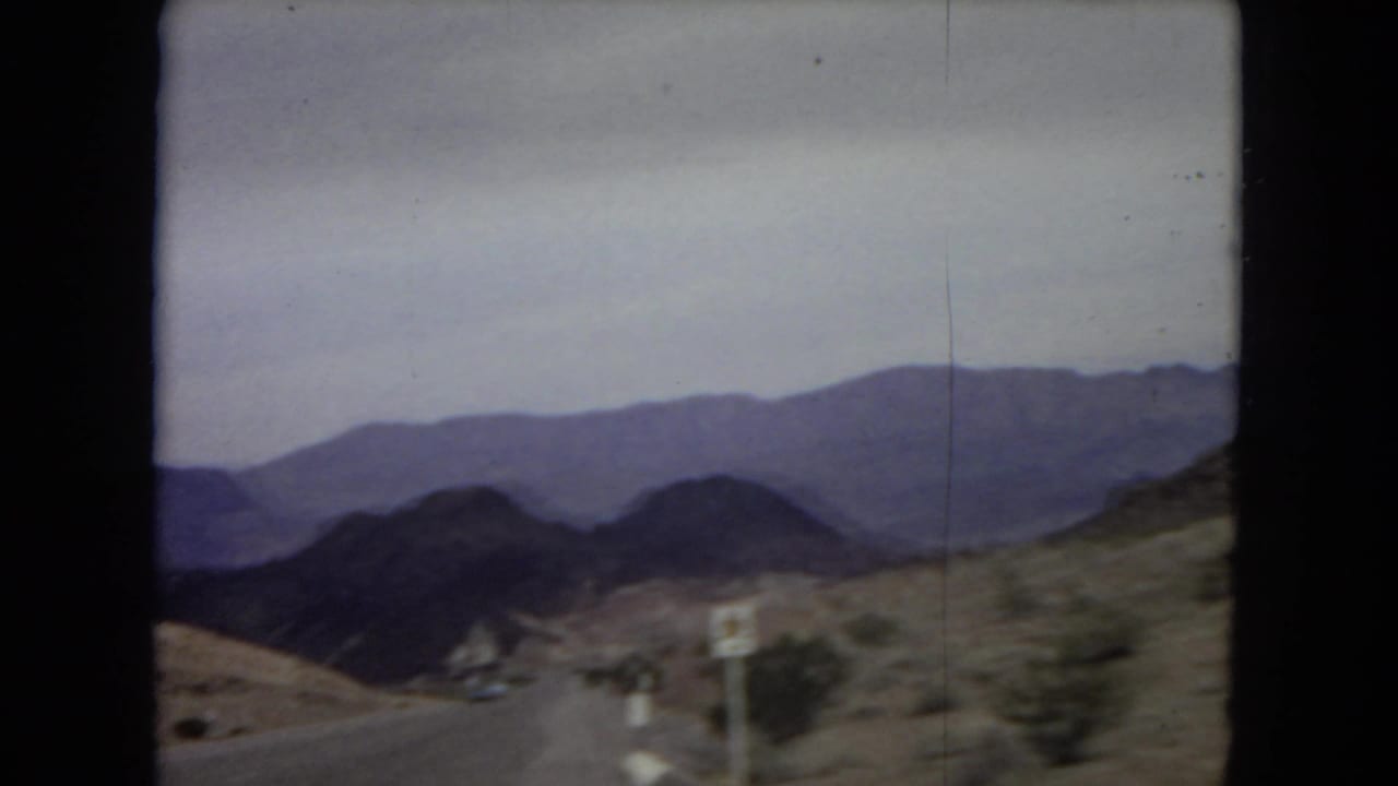 1967 8mm frame from a moving vehicle showing a desert road, rugged mountains, and a roadside sign associated with the Lake Mead National Recreation Area.