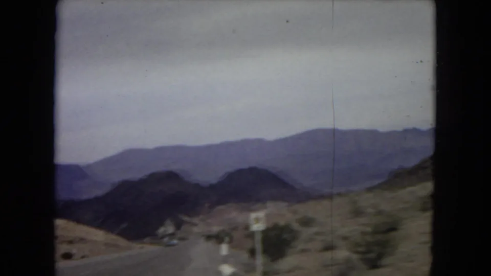 1967 8mm frame from a moving vehicle showing a desert road, rugged mountains, and a roadside sign associated with the Lake Mead National Recreation Area.