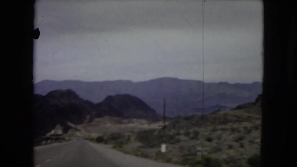 1967 8mm home-movie frame showing a two-lane desert road leading toward dark mountain slopes and distant hazy ridges.