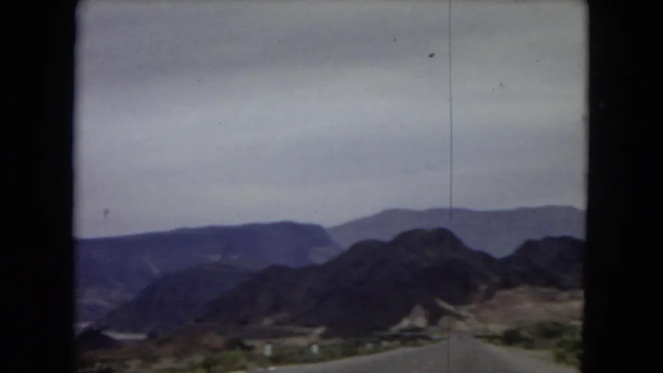 1967 8mm frame showing a desert road descending toward a mountain basin with layered ridges under a pale, hazy sky.