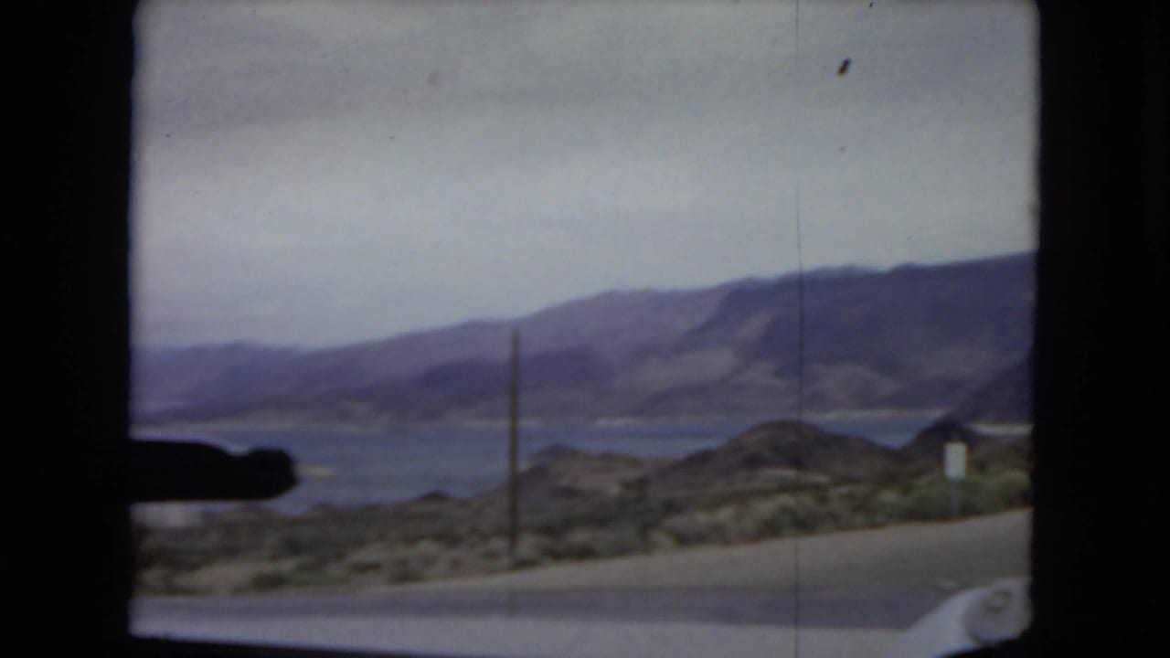 Arizona 1967 travel view through a car windshield, with a road, desert hills, utility pole, mountains, and a large body of water in the distance.