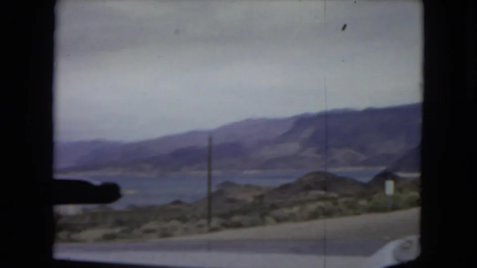 Arizona 1967 travel view through a car windshield, with a road, desert hills, utility pole, mountains, and a large body of water in the distance.