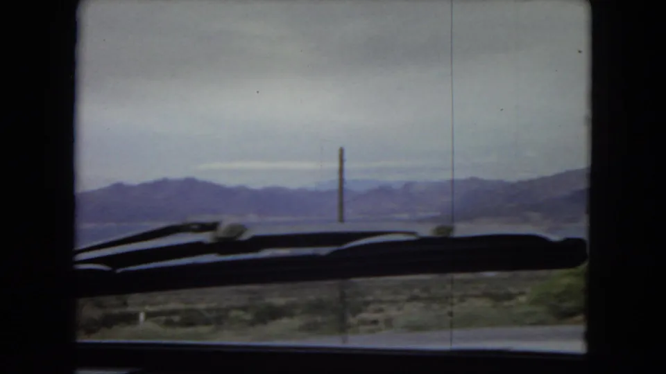 Arizona 1967 home-movie frame from inside an automobile, showing a windshield wiper across the foreground and distant mountains beyond open water.