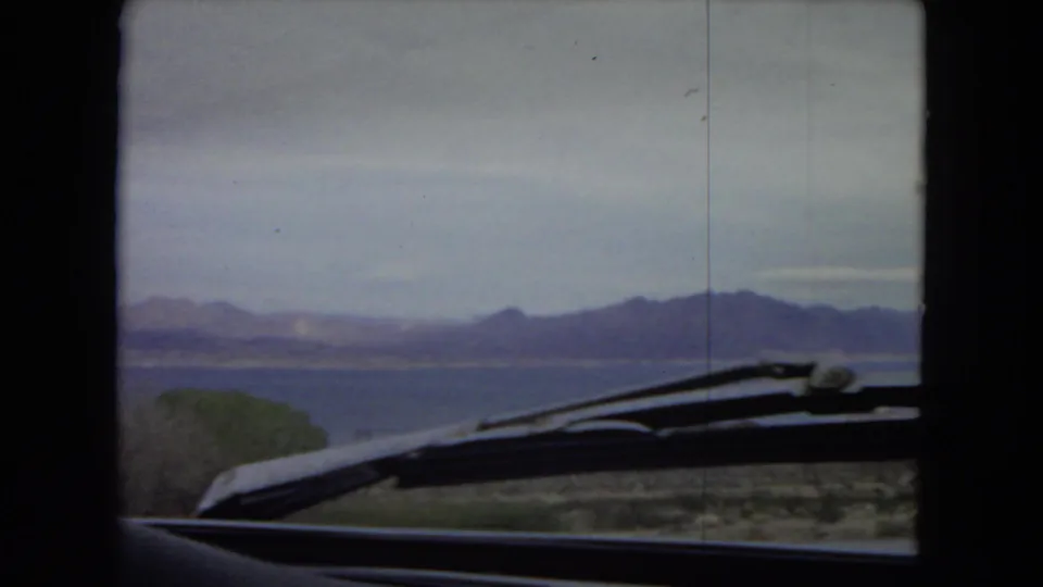 Arizona 1967 automobile-view landscape with the windshield wiper and dashboard in the foreground, open water beyond the road, and low mountain ranges in the distance.