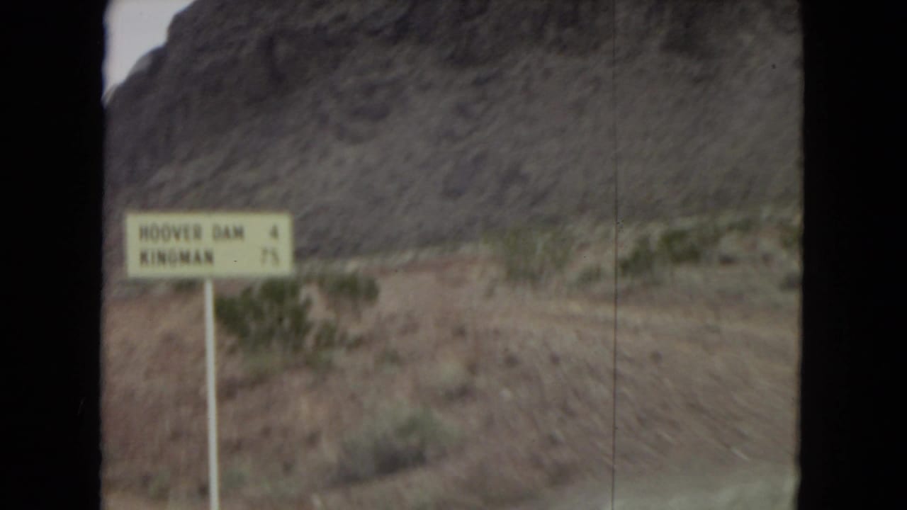 1967 8mm home-movie frame of a yellow roadside direction sign near dry scrub and rocky hills in the American Southwest, seen through motion blur from a passing vehicle.