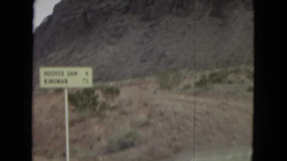 1967 8mm home-movie frame of a yellow roadside direction sign near dry scrub and rocky hills in the American Southwest, seen through motion blur from a passing vehicle.