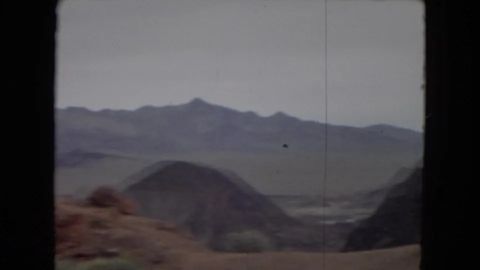 A wide desert valley with low rounded hills and distant mountain ridges appears in a 1967 8mm travel view shot from a moving car.