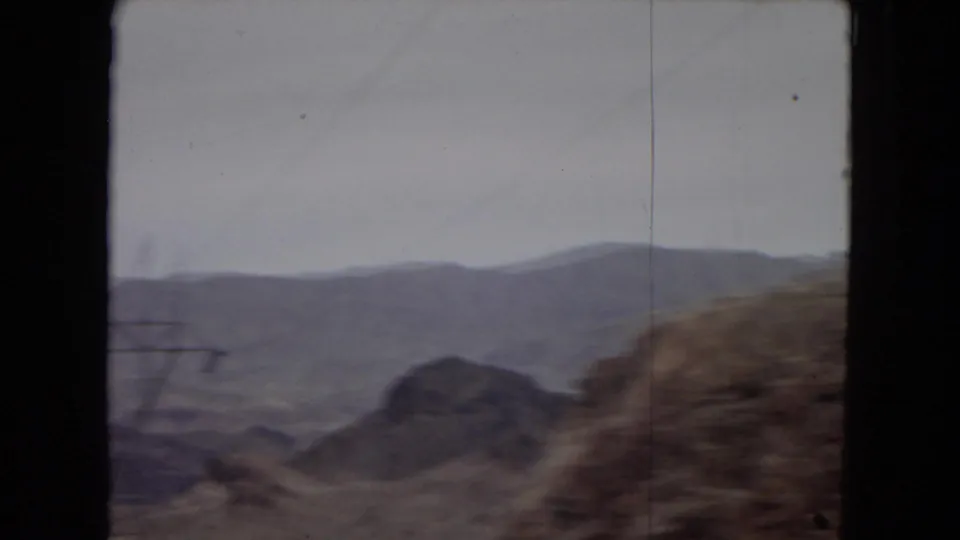 Muted 8mm view of distant Southwestern mountains and rocky roadside terrain passing the camera during a car ride.
