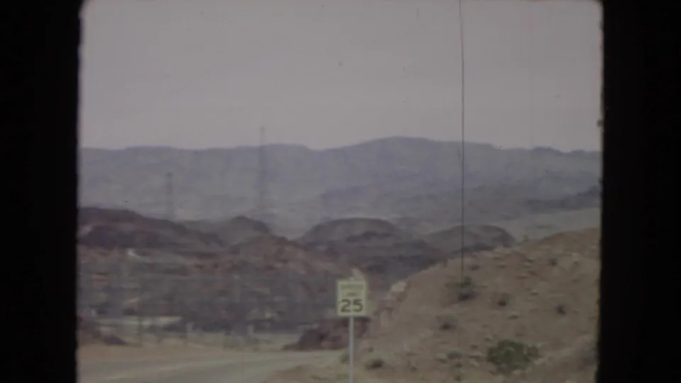 Roadside 1967 home-movie frame showing a paved route through desert hills, a visible speed-limit sign, and hazy mountain ridges beyond.