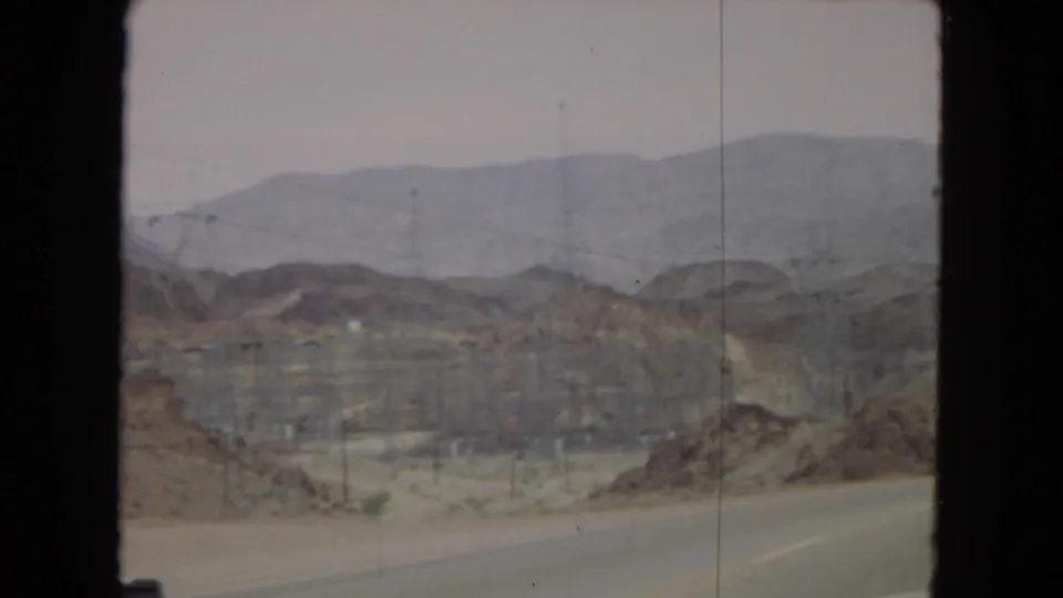 Moving-vehicle view of a desert road near Hoover Dam area metadata, with transmission towers, utility lines, low hills, and distant mountains.