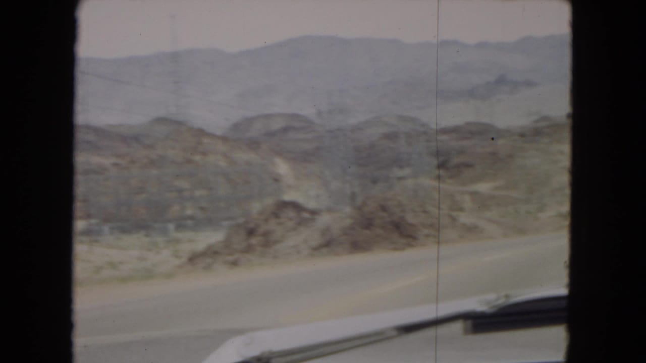 1967 Arizona home-movie frame showing a paved desert road from a car-window viewpoint, with rocky arid hills, distant mountain ridges, hazy sky, and part of the vehicle visible at the bottom edge.
