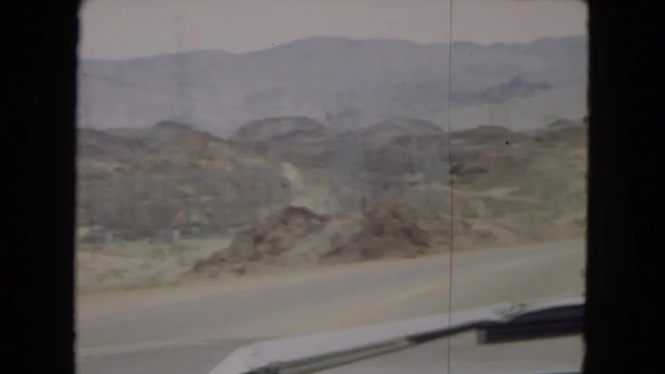 1967 Arizona home-movie frame showing a paved desert road from a car-window viewpoint, with rocky arid hills, distant mountain ridges, hazy sky, and part of the vehicle visible at the bottom edge.