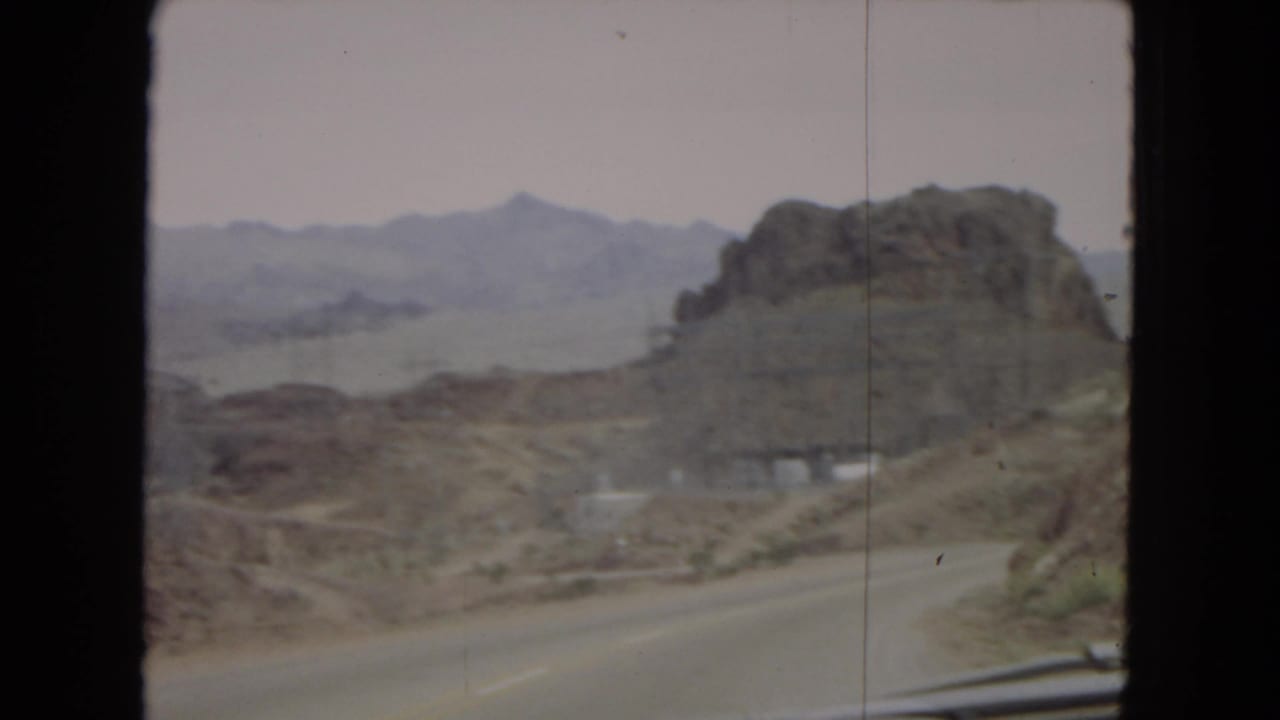 1967 Arizona road-trip frame showing a curving desert highway near a large rocky outcrop, with a white car visible on the roadway and mountain ridges fading into the hazy background.