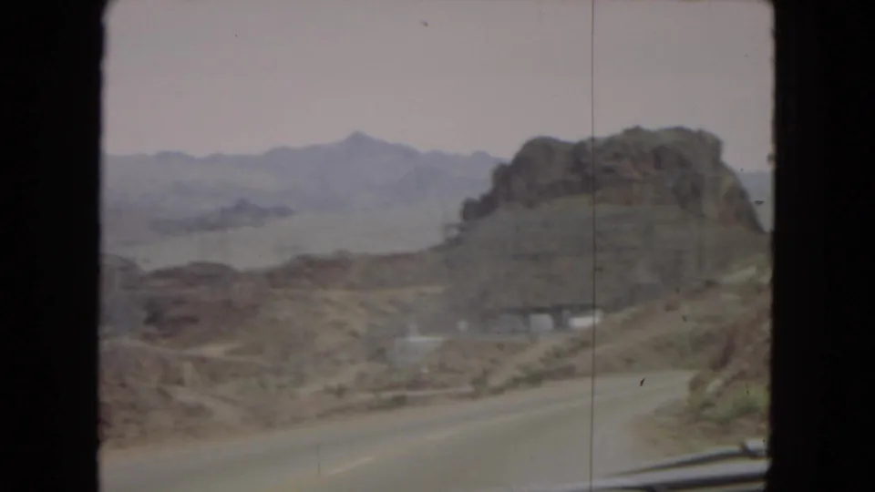1967 Arizona road-trip frame showing a curving desert highway near a large rocky outcrop, with a white car visible on the roadway and mountain ridges fading into the hazy background.