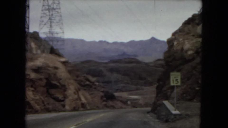 1967 Arizona home-movie frame showing a road descending through rocky desert terrain, with a lattice utility tower at left, a 15 mph speed-limit sign at right, and distant mountains beyond.