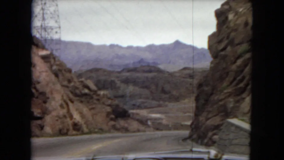 1967 Arizona road-trip frame showing a paved road passing between rocky desert cuts, with a low roadside barrier, distant ridges, pale sky, and the car-window viewpoint still evident.