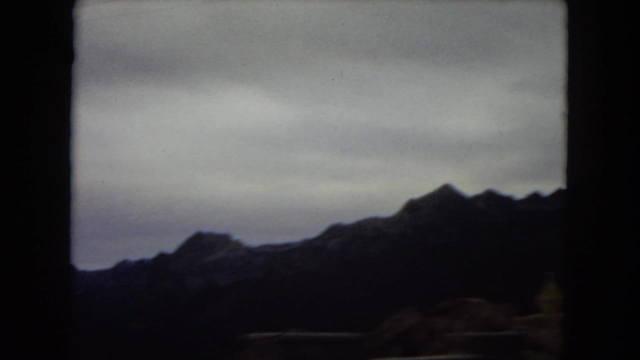 1967 U.S. home-movie frame, reportedly Arizona, showing distant rugged mountain silhouettes through a dark vehicle window under a pale overcast sky.