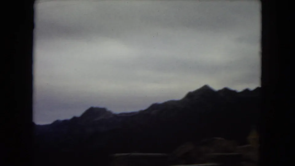 1967 U.S. home-movie frame, reportedly Arizona, showing distant rugged mountain silhouettes through a dark vehicle window under a pale overcast sky.