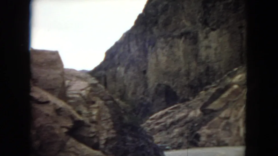 Rugged cliff face and broken rocky ground viewed from a moving vehicle in archival 8mm footage, with bright sky and dark window framing at the edges.