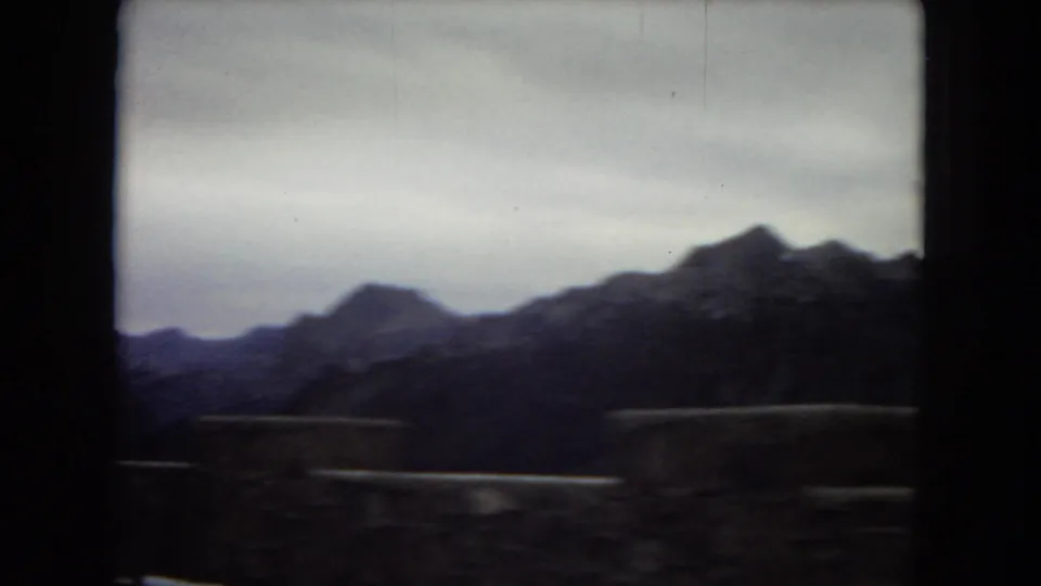 Distant rugged mountain ridge seen through a dark car-window frame, with low roadside foreground shapes and an overcast sky in 1960s home-movie footage.