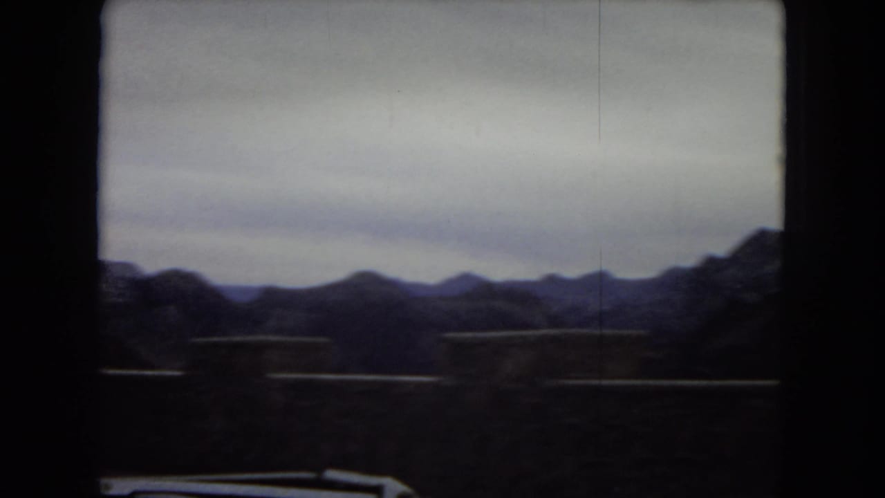 1967 8mm road-trip frame looking across a highway barrier toward dark desert mountains under a pale sky.