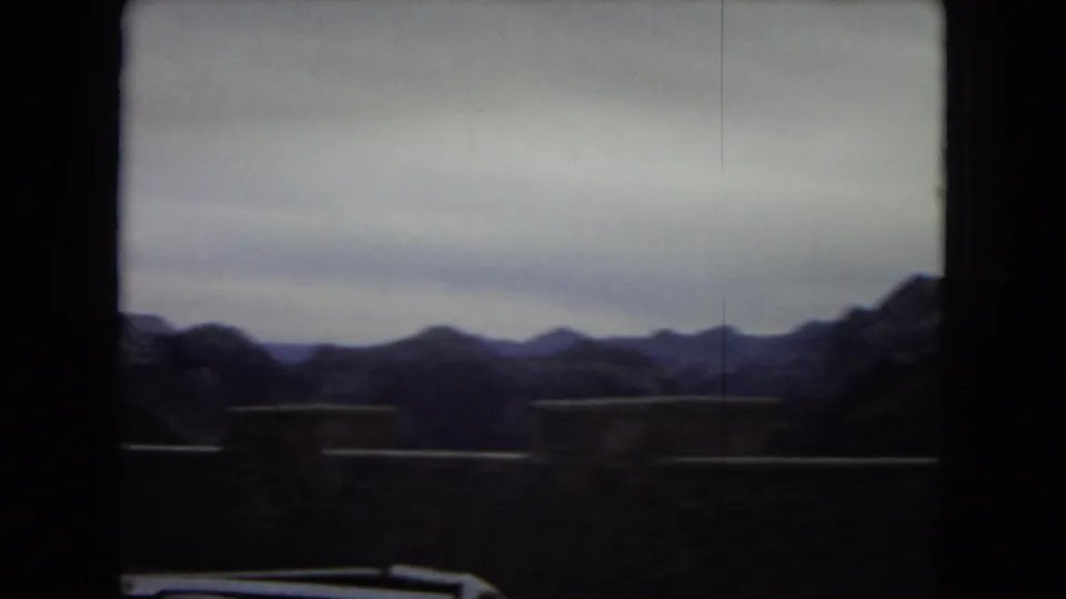 1967 8mm road-trip frame looking across a highway barrier toward dark desert mountains under a pale sky.