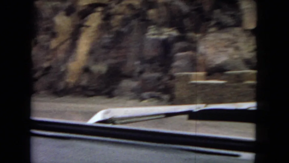 Moving-car view of rocky desert roadside terrain with the car hood and windshield wipers visible in the foreground.