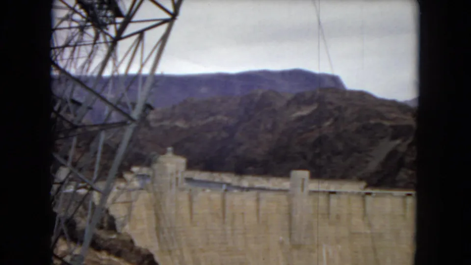 1967 home-movie view of Hoover Dam framed beside a metal utility tower, with dry hills rising behind the concrete structure.