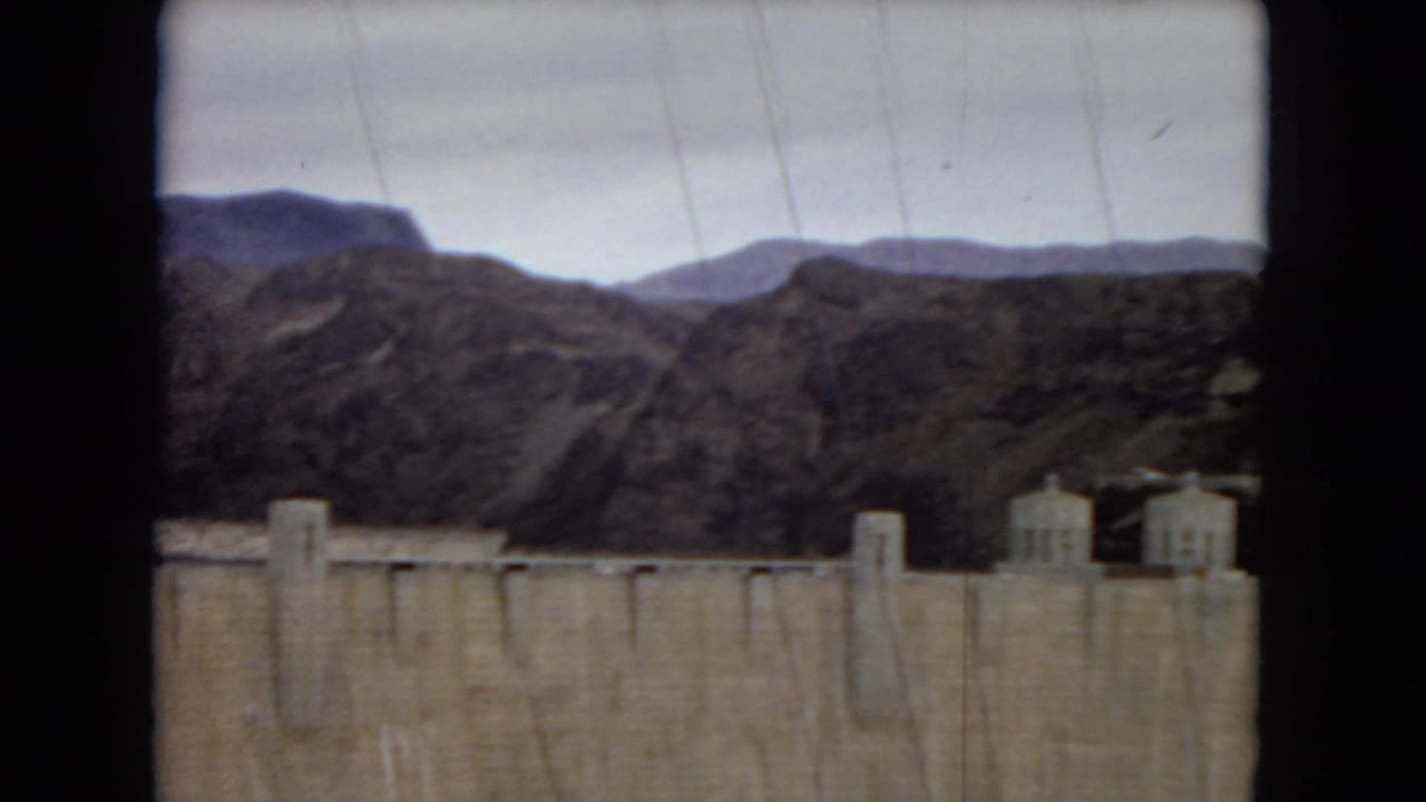 1967 8mm view of Hoover Dam, showing the concrete dam face, small power plant structures, rocky desert mountains, and cloudy sky.