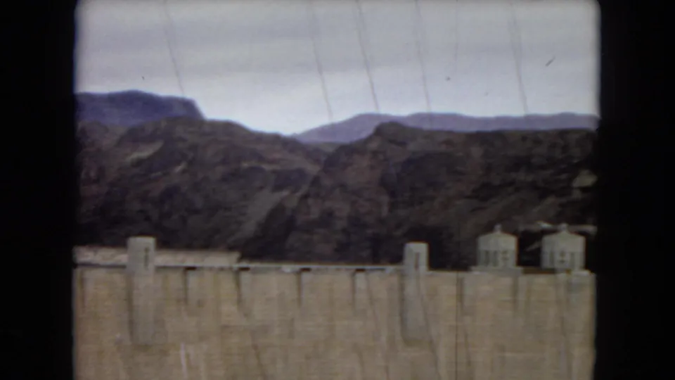 1967 8mm view of Hoover Dam, showing the concrete dam face, small power plant structures, rocky desert mountains, and cloudy sky.