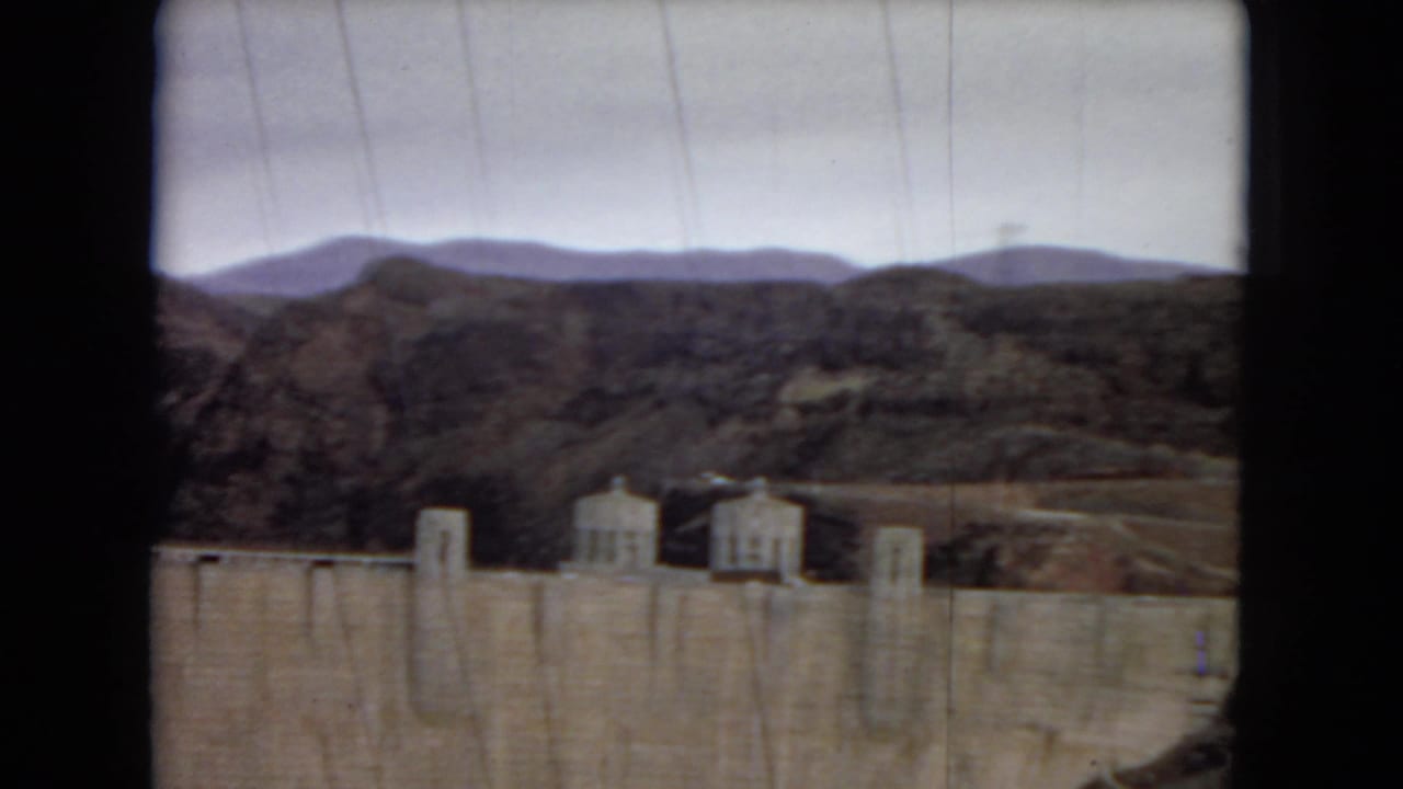 Concrete dam and power plant structures with hills in the background.