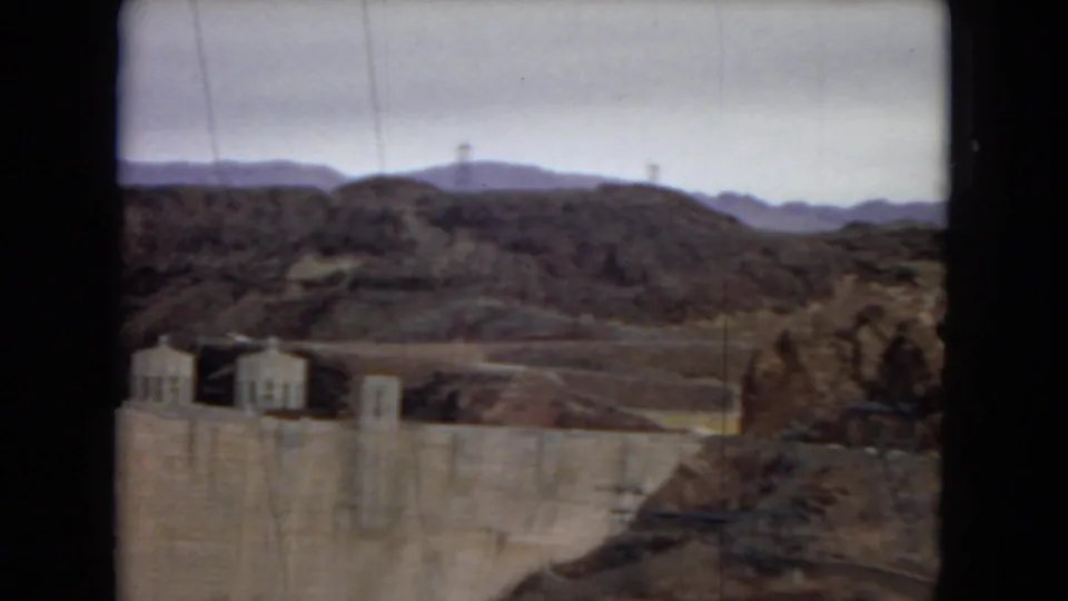 1967 home-movie frame showing part of Hoover Dam at the lower edge, with rocky desert terrain and mountain ridges beyond.