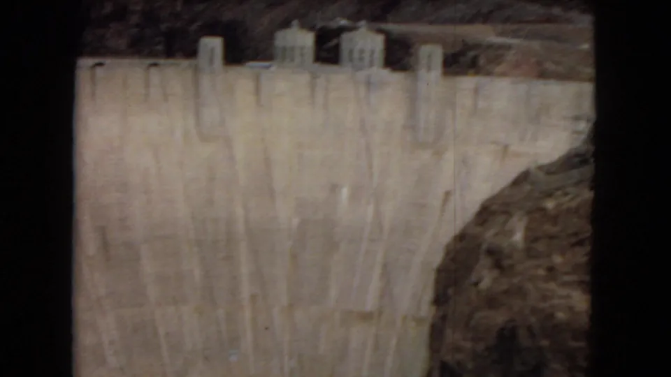 8mm frame of Hoover Dam's curved concrete face with power plant structures above and a rocky canyon wall along the side.