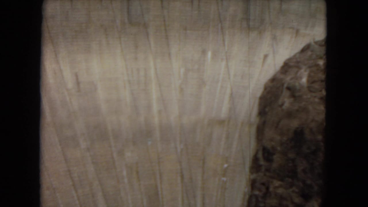 1967 Arizona home-movie frame showing the upper curved face of Glen Canyon Dam with vertical marks and dark canyon rock at the side.