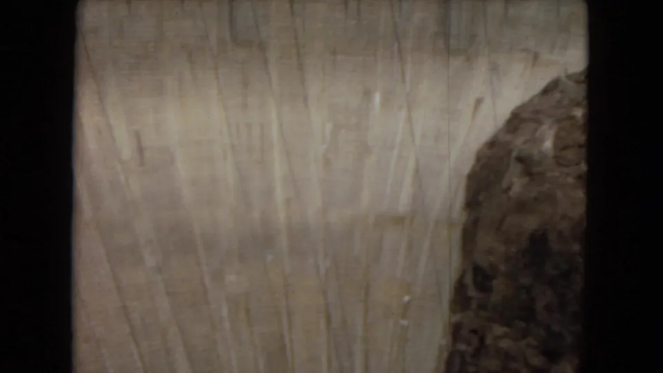1967 Arizona home-movie frame showing the upper curved face of Glen Canyon Dam with vertical marks and dark canyon rock at the side.