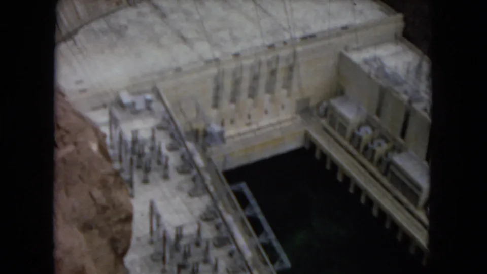 1967 Arizona home-movie frame showing Glen Canyon Dam's power plant infrastructure, dark water, and the massive concrete wall above.