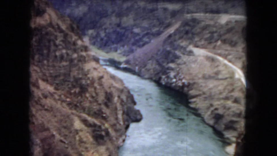 1967 8mm home-movie frame showing a winding river cutting through a rugged Arizona canyon, with dry slopes, rocky cliffs, and a pale road or path near the upper right.