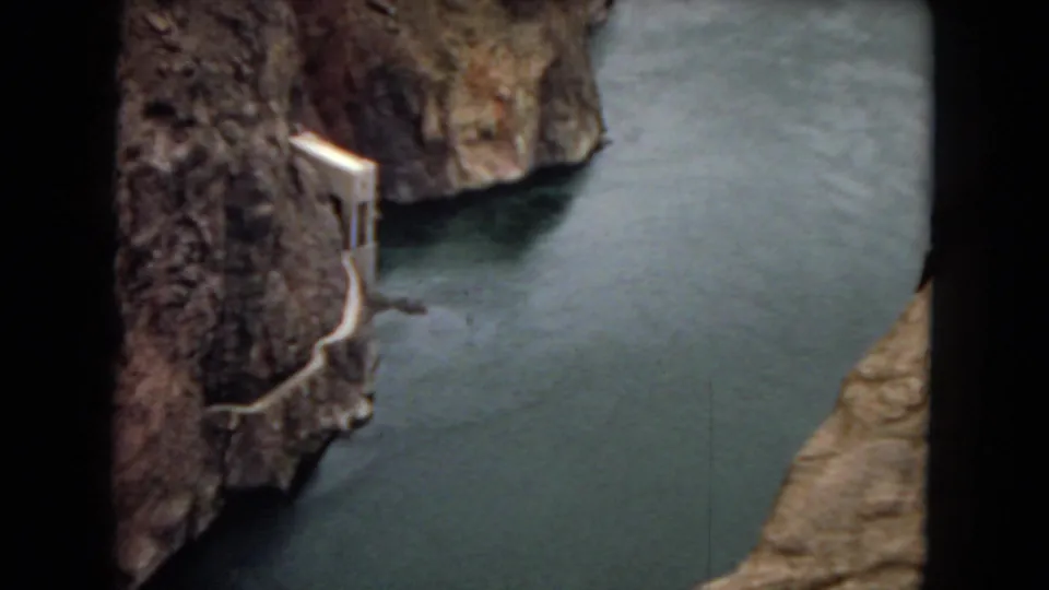 Closer 1967 8mm view of an Arizona canyon river, with a steep rock face, dark green water, and a small concrete-looking structure near the shoreline.