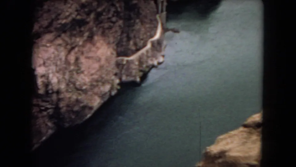 1967 8mm home-movie frame showing a wider Arizona canyon river view, including steep rock walls, a broad water channel, and a small dam-like feature at the left edge.